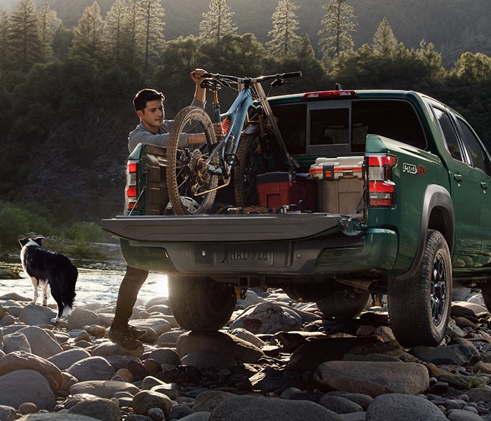 2024 Nissan Frontier parked on a rocky stream shore; the driver is adjusting a bicycle and cargo in the back, while his dog is looking at the stream