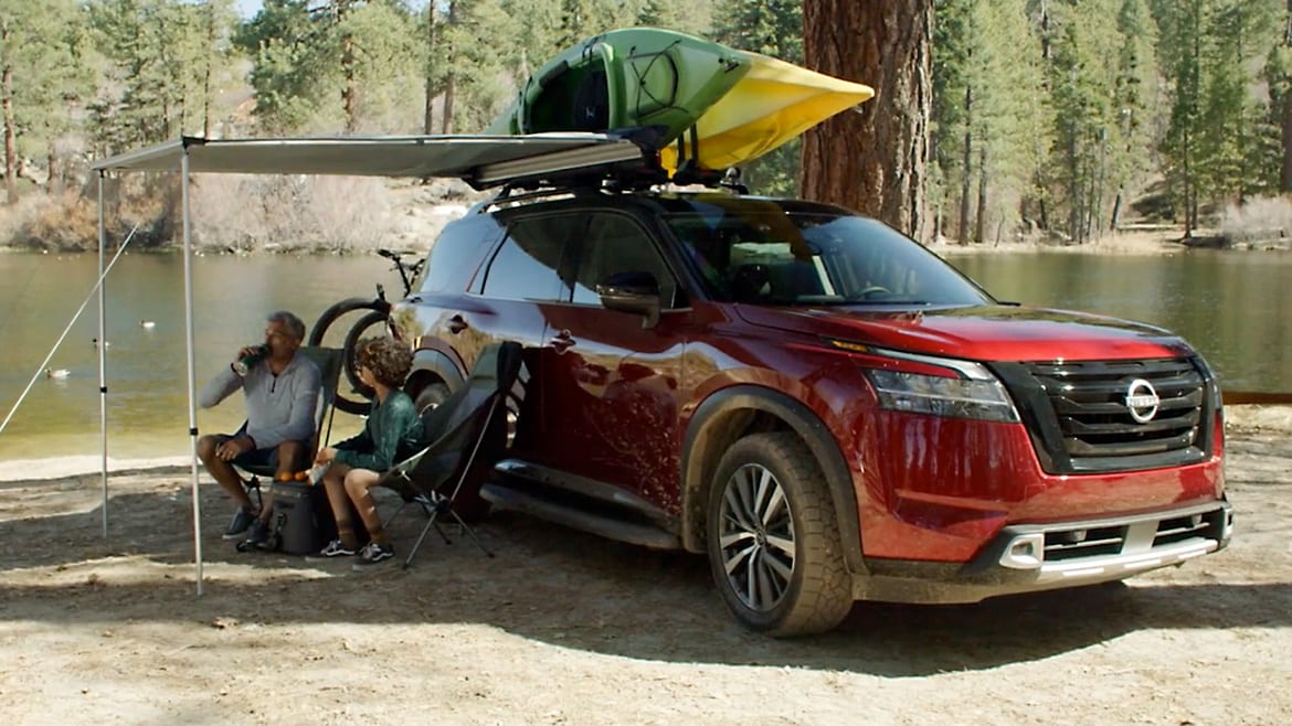 2024 Nissan Pathfinder at the water's edge, with kayaks on top, and a retractable shade over two people in camping chairs.
