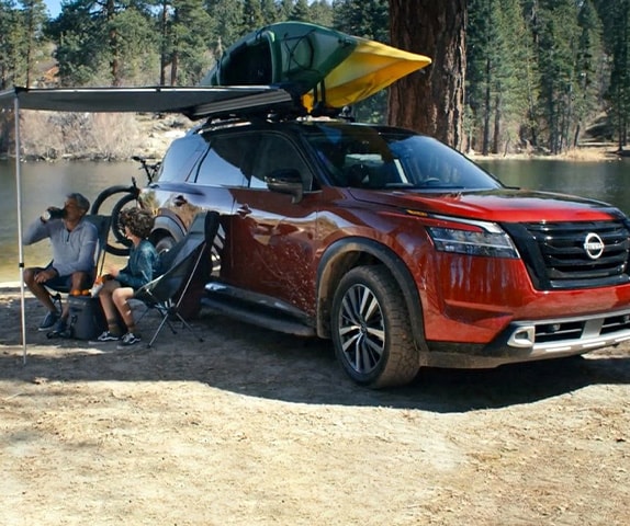 2024 Nissan Pathfinder at the water's edge, with kayaks on top, and a retractable shade over two people in camping chairs.