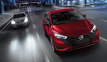 2023 Nissan Versa in Scarlet Ember Tintcoat shown driving downtown at night.